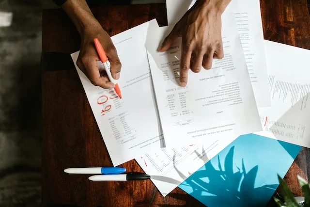Person reviewing and marking a purchase order document with a red pen, highlighting important details. Purchase order paperwork, a blue folder, and pens on a wooden desk. Learn about purchase order numbers and how to create one.